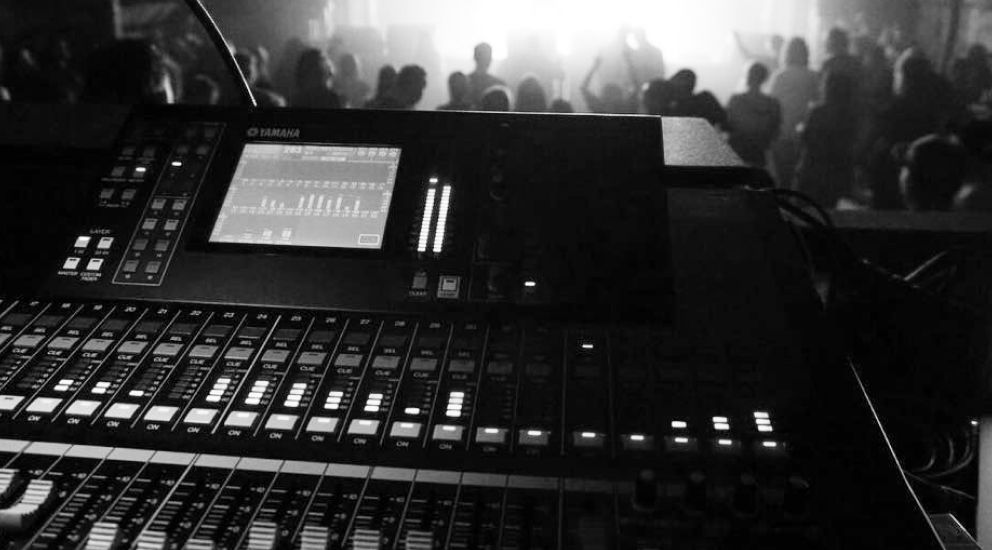 Tonmischpult mit beleuchteten Bildschirmen und Schiebereglern in einem dunklen Raum mit einem unscharfen Publikum im Hintergrund.